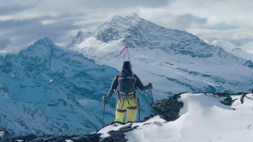 A mountaineer stands atop a mountain ridge and gazes down a snowy mountain valley below