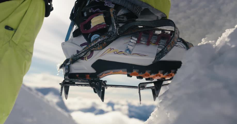 Person wears climbing shoes with metal spike to climb ice with mountains in the background