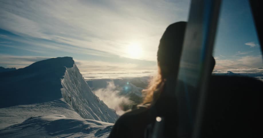 A woman with skis on her back stands on top of a mountain and looks at the landscape