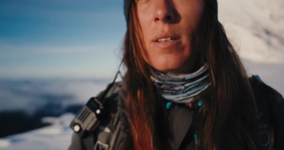 A female mountaineer hikes across a snow mountain ridge before stopping in front of the camera and gazing into the distance