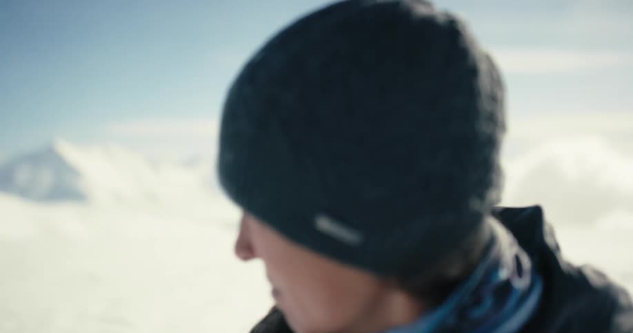 A female mountaineer standing in a field of snow on a snowy day stares into the camera before turning to look at the distant mountains