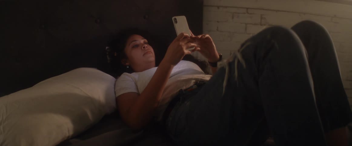 A hispanic woman views media on her smartphone while lying in a bed in a dimly lit room