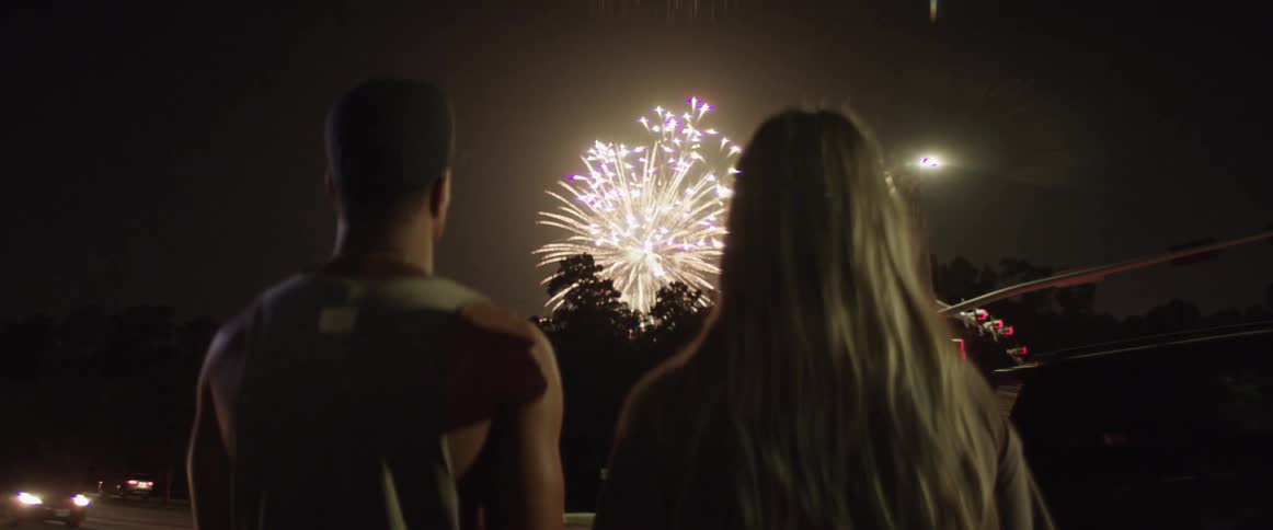 Male and female stand along the side of the road watching fireworks emerge over the trees as car pass by on the roadway