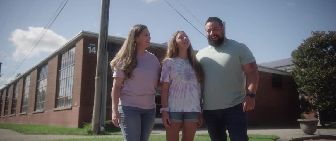 A family laughs while standing together outside of a building on a sunny day