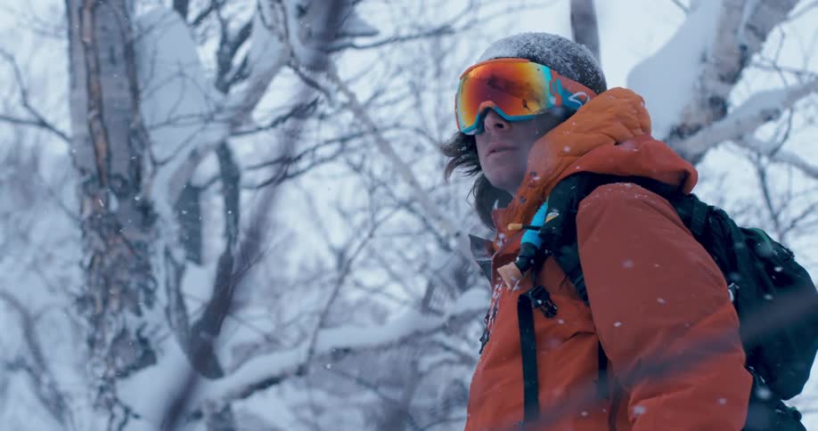 A man wearing an orange coat and ski goggles looks around as the snow falls