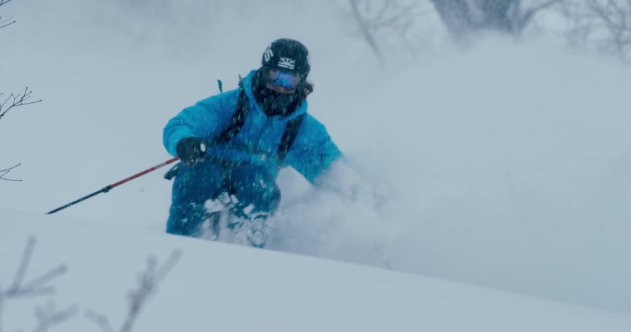 A skier skis down a mountain slope covered in leafless trees as heavy snow falls from the sky