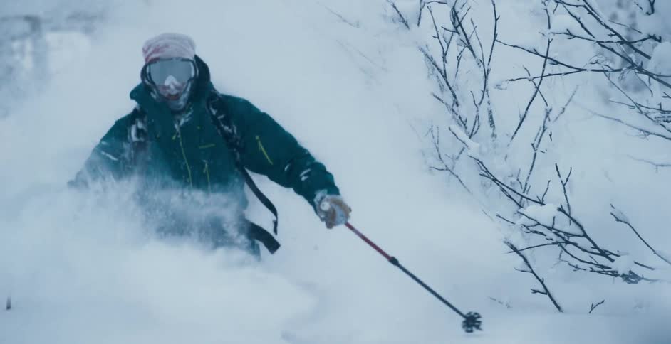 A person with a coat gloves and goggles skiing down a snowy slope with trees