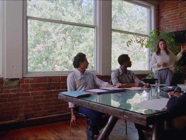 A group of people in an office with paperwork at a table listening to a female speaker