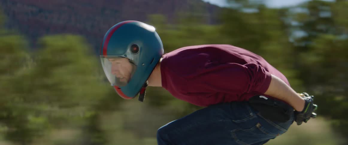 Three men with helmets ride skateboards at high speed on a country road with mountains in the background