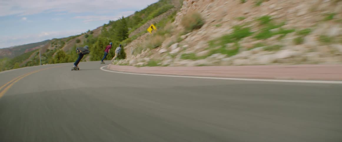 Three people skateboard down a blind curve road beside a large mountain