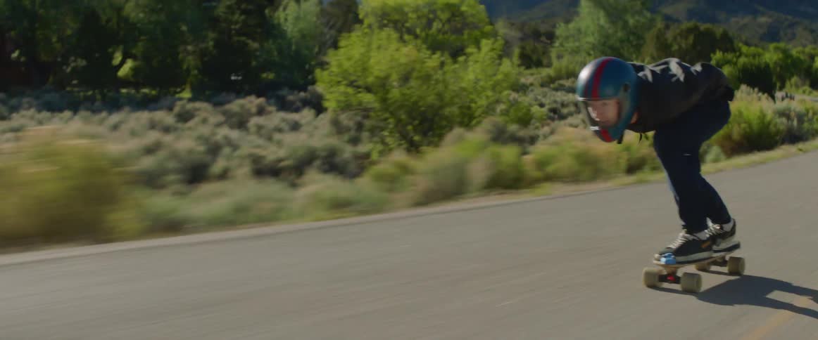 Person skateboards down winding highway past mountains and scrub