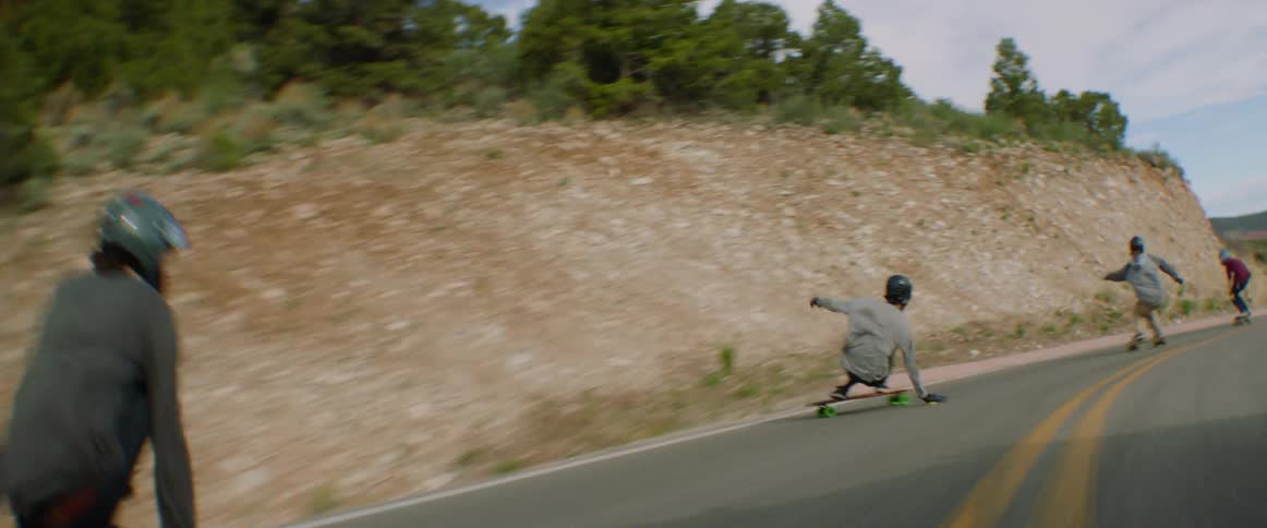 A group of skateboarders travel along a countryside road at high speed on a sunny day