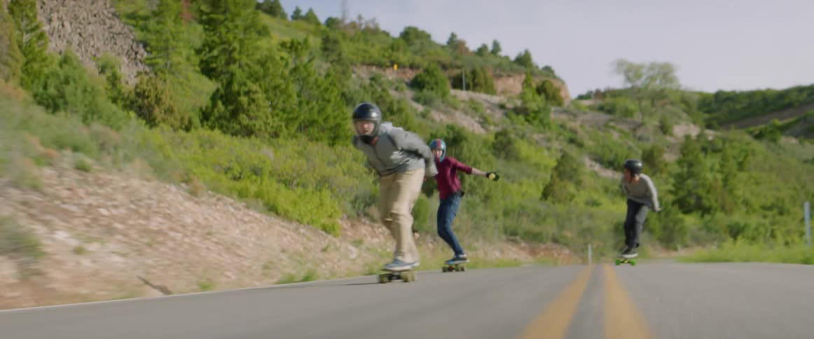 A skateboarder riding at high speed down a road through the countryside looks back to gesture at the skaters following behind