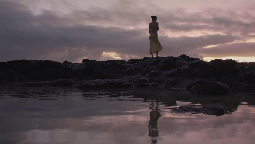 A young woman stretches while standing on a rock formation on a lake in the evening