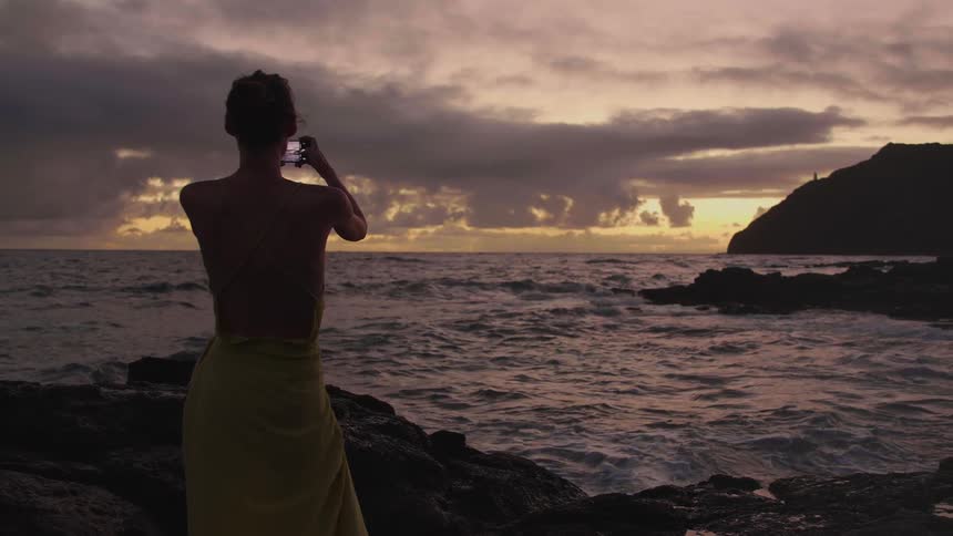 Woman with a dress takes photographs of the sea and the promontory overlooking the bay at dusk