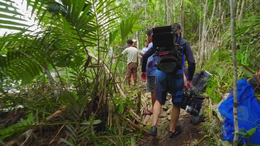 A man carries large filming equipment through a forest to the shore of a lake where a group of people await him