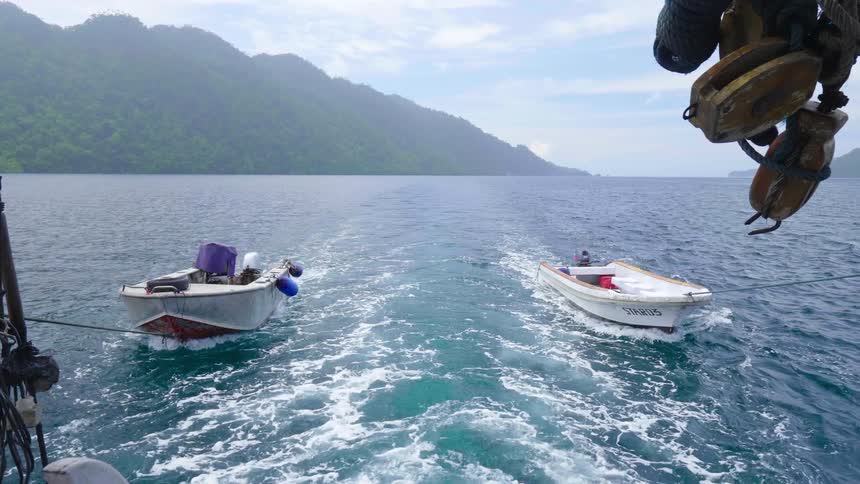 A pair of boats are towed behind a large vessel across the ocean on a sunny day