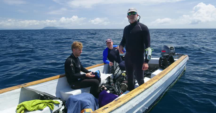 Three white men in diving suits are sitting and standing in a white boat in the middle of a blue ocean under a blue cloudy sky