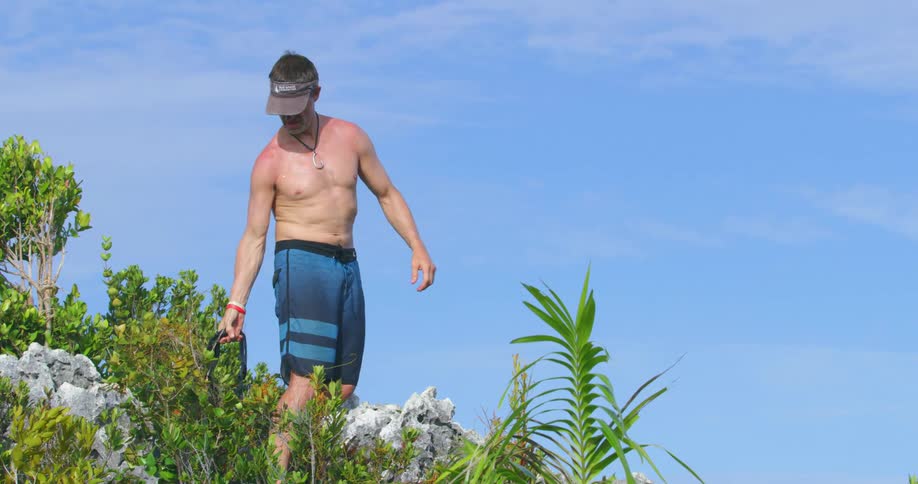A man stands alone and takes a backpack off of his back and wearing swim trunks