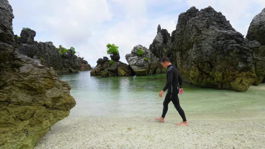 A man in a wet suit walks along a beach surrounded by large rock formations