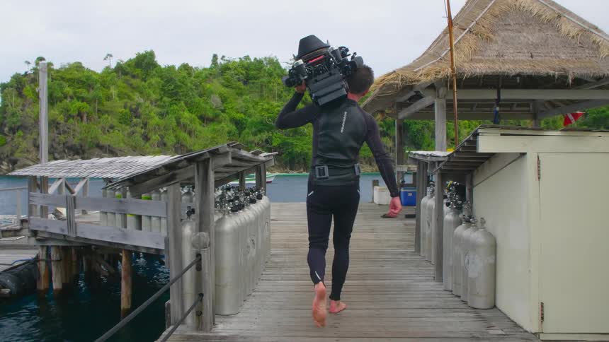 A man walking on a pier holding a camera on his shoulders with the ocean and a boat in the background