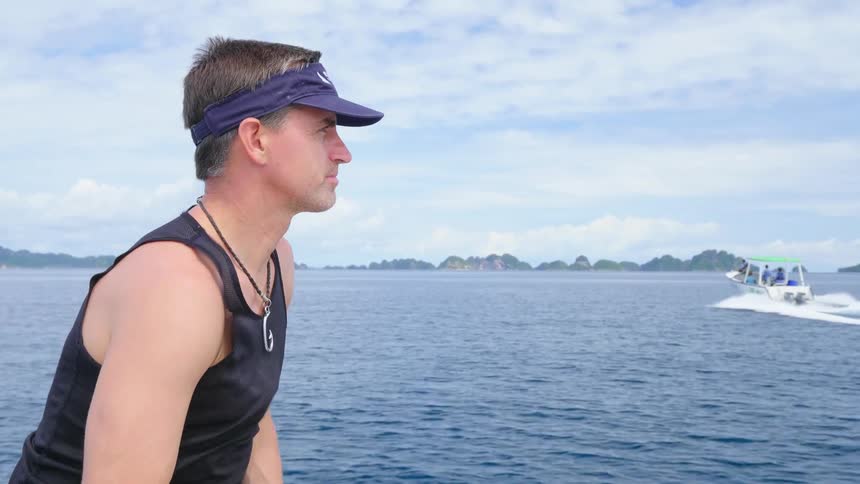 A man wearing a sleeveless shirt and visor watches a small boat traveling across the water