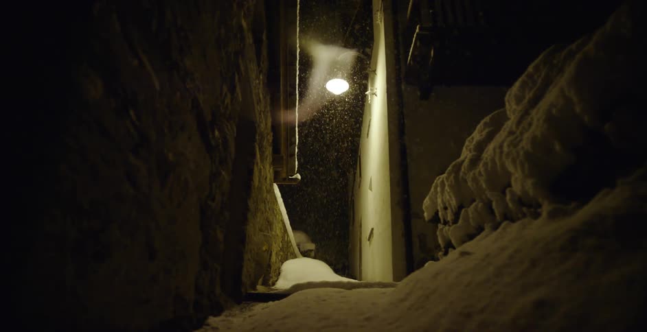 Snow falls in a narrow alleyway at night