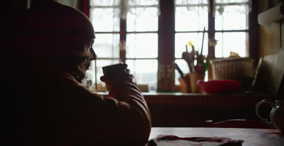 A person sits at a kitchen table in front of a large window drinking a hot steaming beverage from a mug