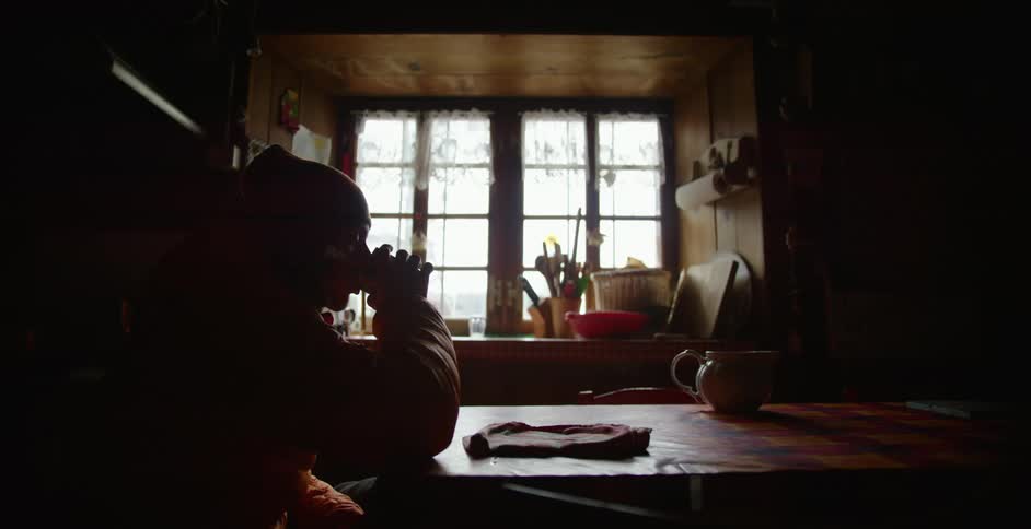 A man wears a knit hat and coat while sitting at his kitchen table drinking coffee