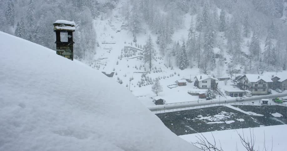 Snow falling over a neighborhood with cars driving down a street and a hilly forest behind houses