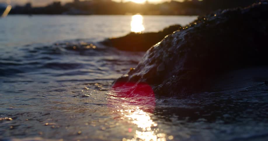 Waves splash against rocky shore as sun shines past city skyline to reflect off ocean and wet sand