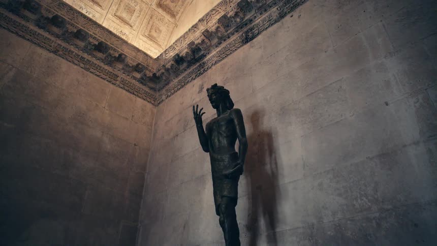 Bronze statue stands on pedestal against marble tiled wall under ornate ceiling