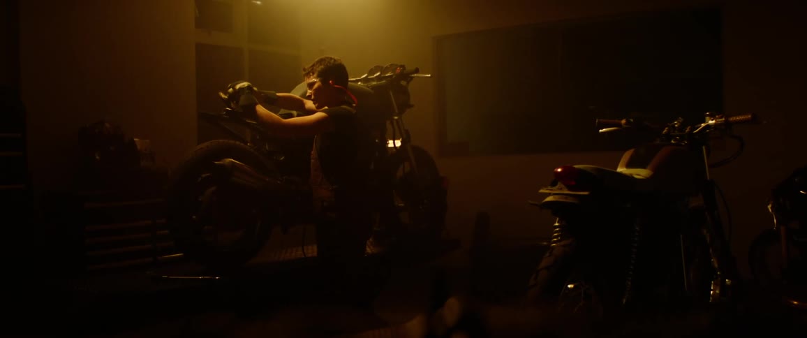 A woman in a dimly lit garage uses an angle grinder on the chassis of a motorcycle