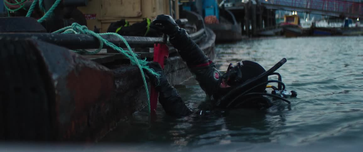 A person wearing scuba gear floats in the water beside a boat holding a tool attempting to fix something
