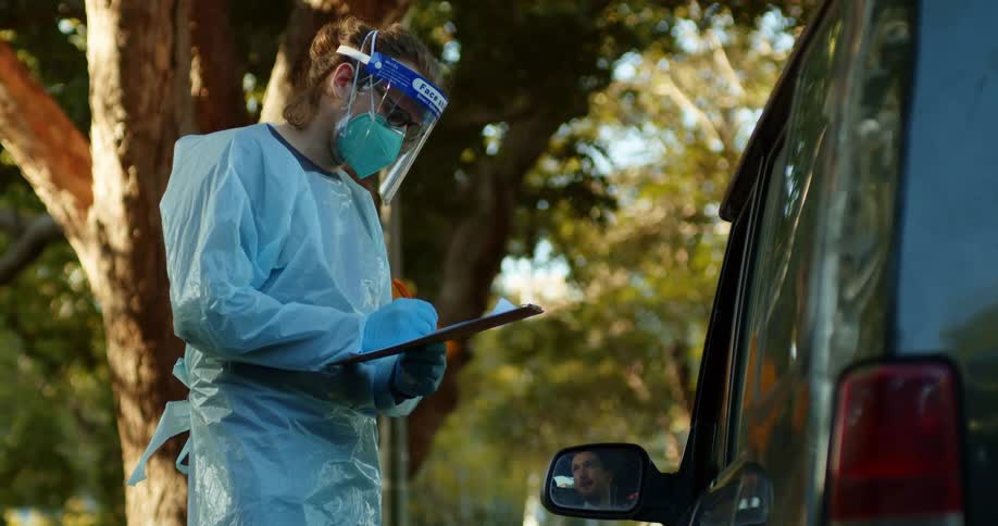 A medical professional wears full protective gear holding a clipboard next to a vehicle at drive through COVID test clinic