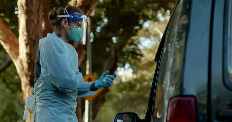 A nurse wearing full medical protective gear stands next to a vehicle and the drivers side door A medical professional wears full protective gear holding a clipboard next to a vehicle at a drive through COVID test clinic