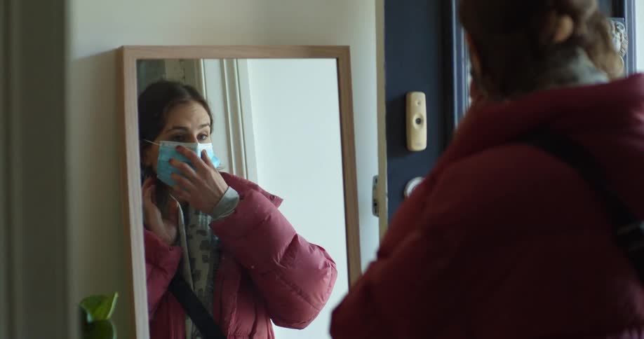 A woman stops in front of a mirror to put on a blue face mask before leaving her house