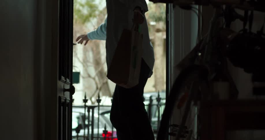 A white man in a long sleeve shirt is opening and closing a front door to a home coming inside holding a bag