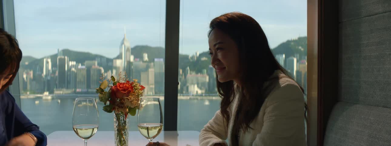 Two women chat and drink wine at a restaurant with views of a river and city skyline