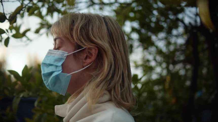 A woman standing outside and putting on a mask with trees the sky and clouds in the background