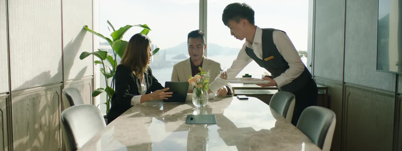 Two people in business attire consult over tablet at table as waiter delivers coffees