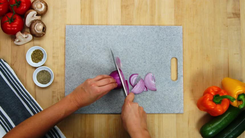 A woman chops a red onion on a gray cutting board with other vegetables spread around her