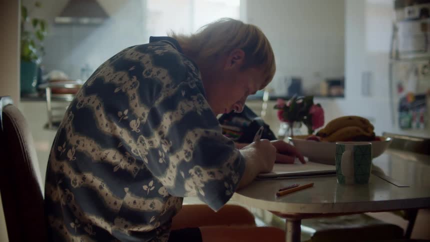 A man writes in a notebook while sitting at a table in a small home kitchen during the day