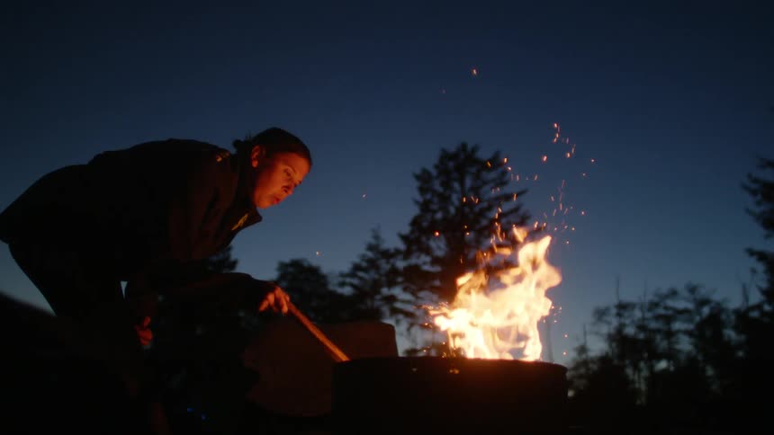 A young woman stirs a fire in a firepit outside