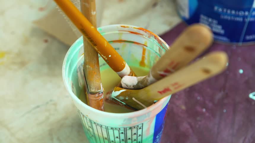 Brushes soak in bucket full of water on paint splattered drop cloth near paint can