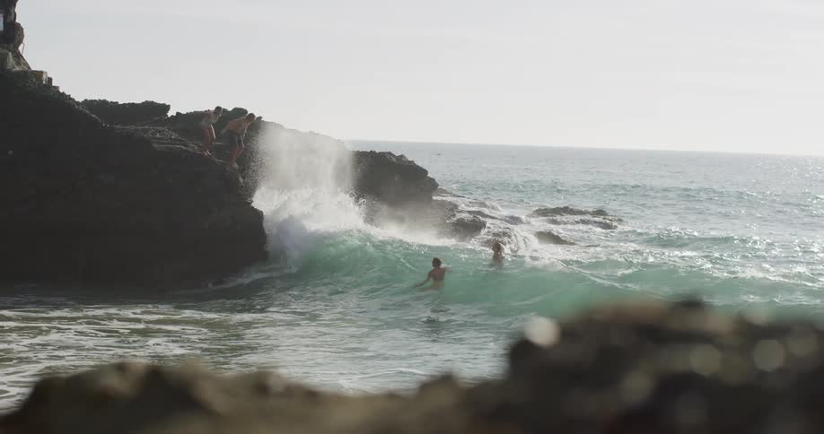 A man and woman in swimsuit jump off a rock formation into the ocean where a pair of swimmers wait for them