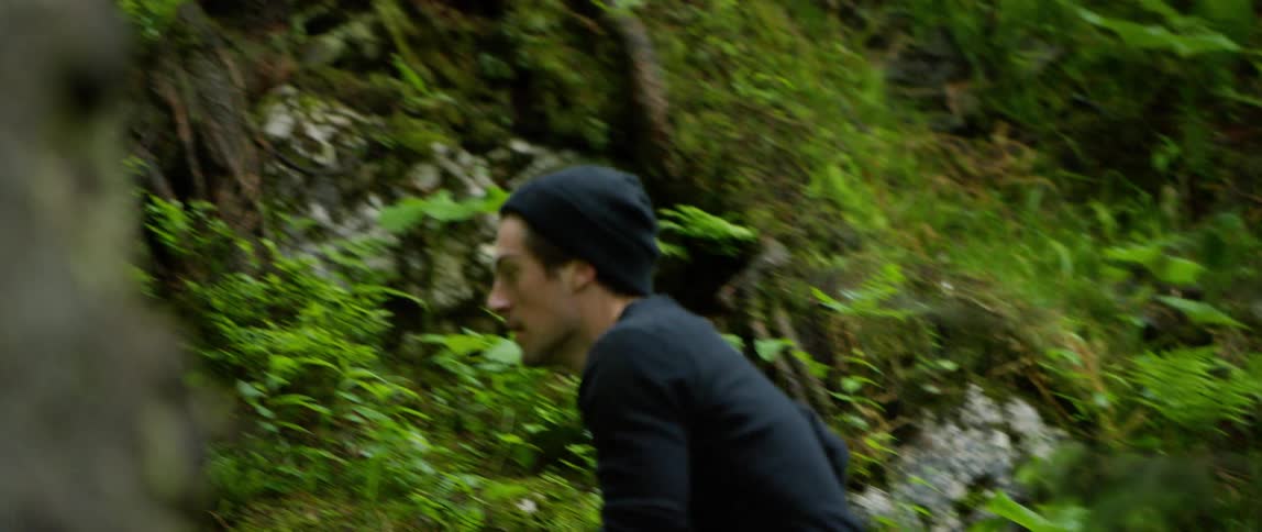 A white man in black clothing and a black hat is running up a steep plant filled forest hill