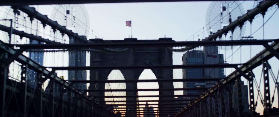 An american flag waves atop the tower of a large bridge in a metropolis in the evening