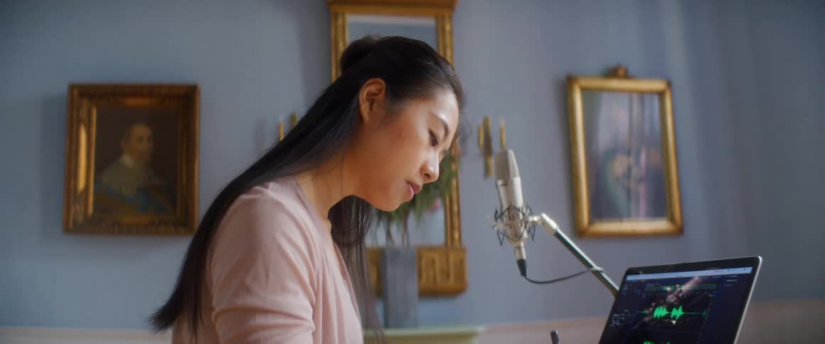 A musician takes notes while standing at her desk in front of a laptop and microphone