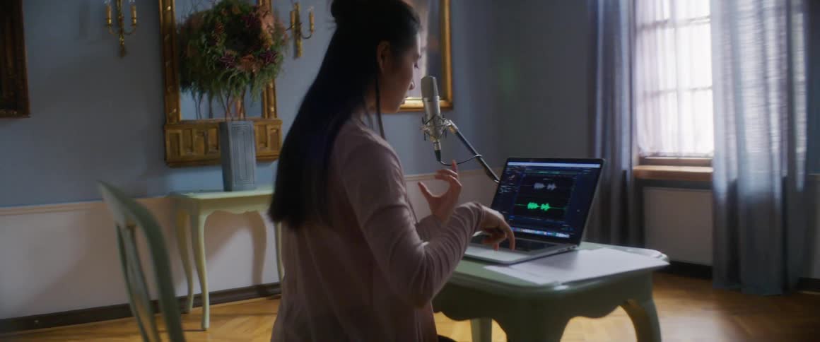 Young woman sits at table with microphone and laptop listening to music
